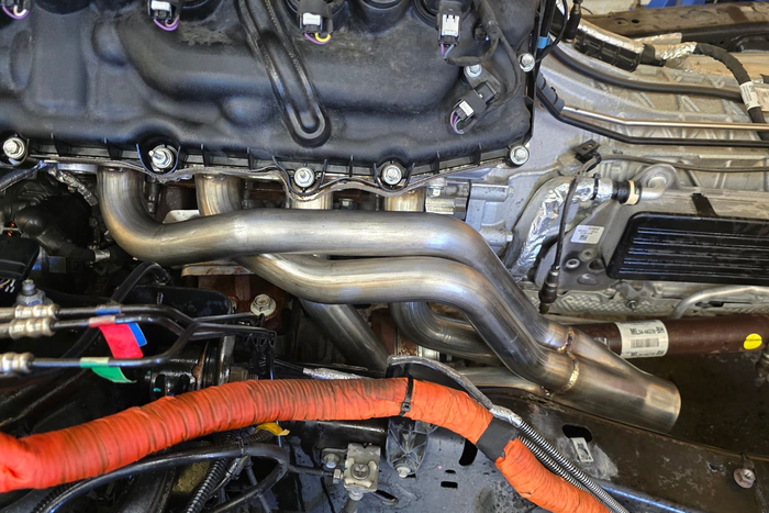 Close-up of a car engine bay with metal pipes, black hoses, and an orange insulated wire harness | Lyons Precision Auto Care