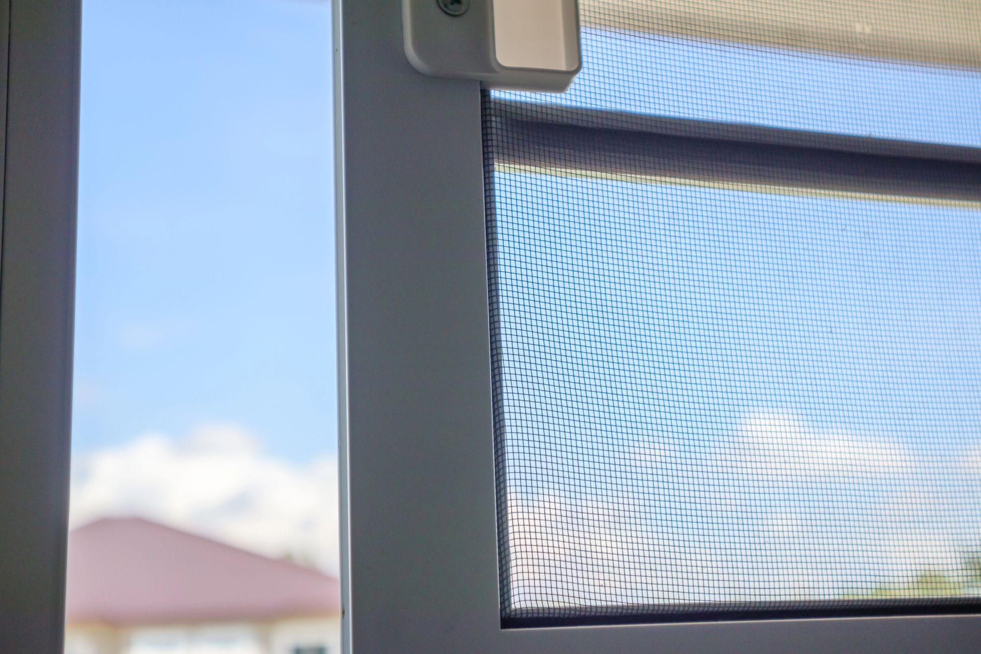 Primo piano di una zanzariera con il bordo strappato, sullo sfondo di un cielo azzurro brillante, con uno scorcio di casa.
