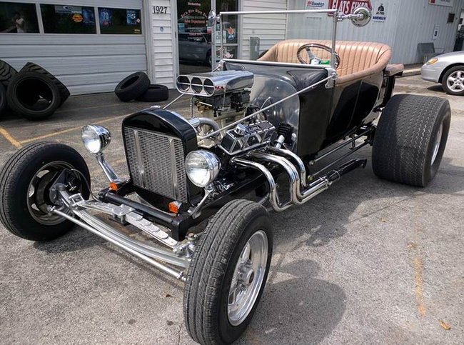 Black hot rod with exposed engine and tan interior parked on a street, front view.
