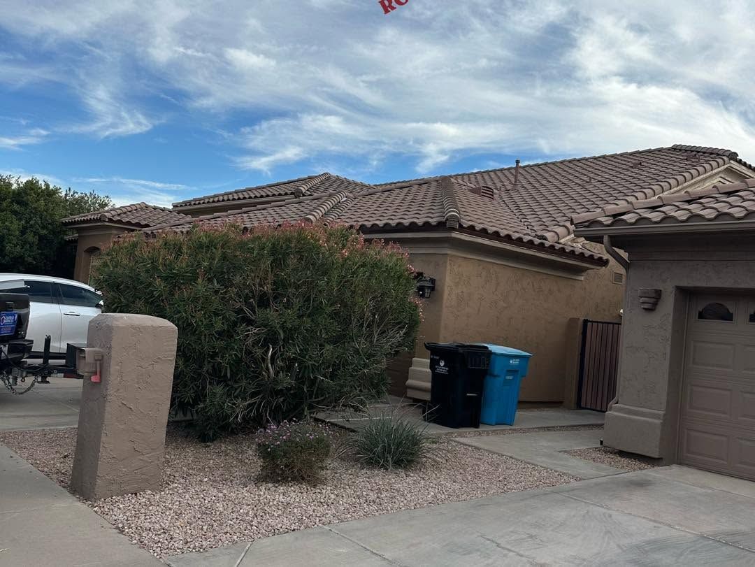 Suburban house exterior with tiled roof, shrubs, driveway, and two trash bins by the garage