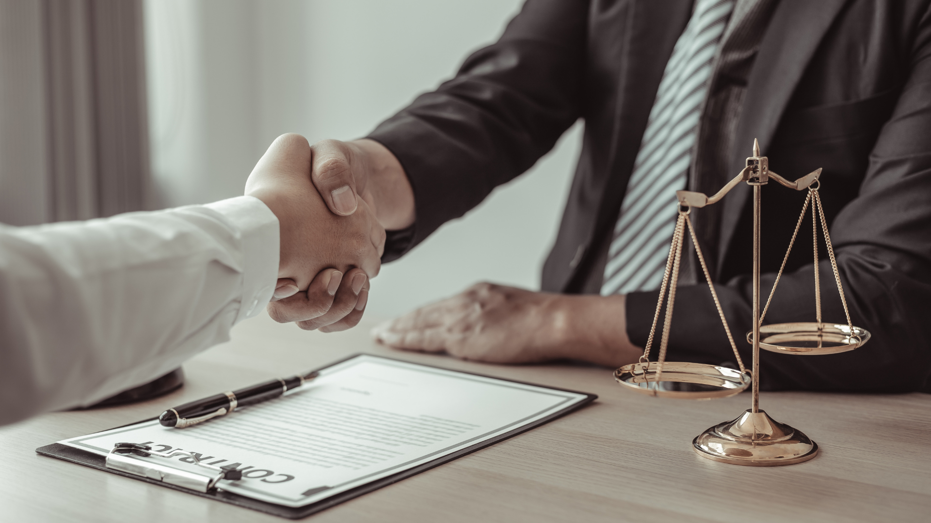 Two people shaking hands over a contract, scales of justice and a pen on the desk.