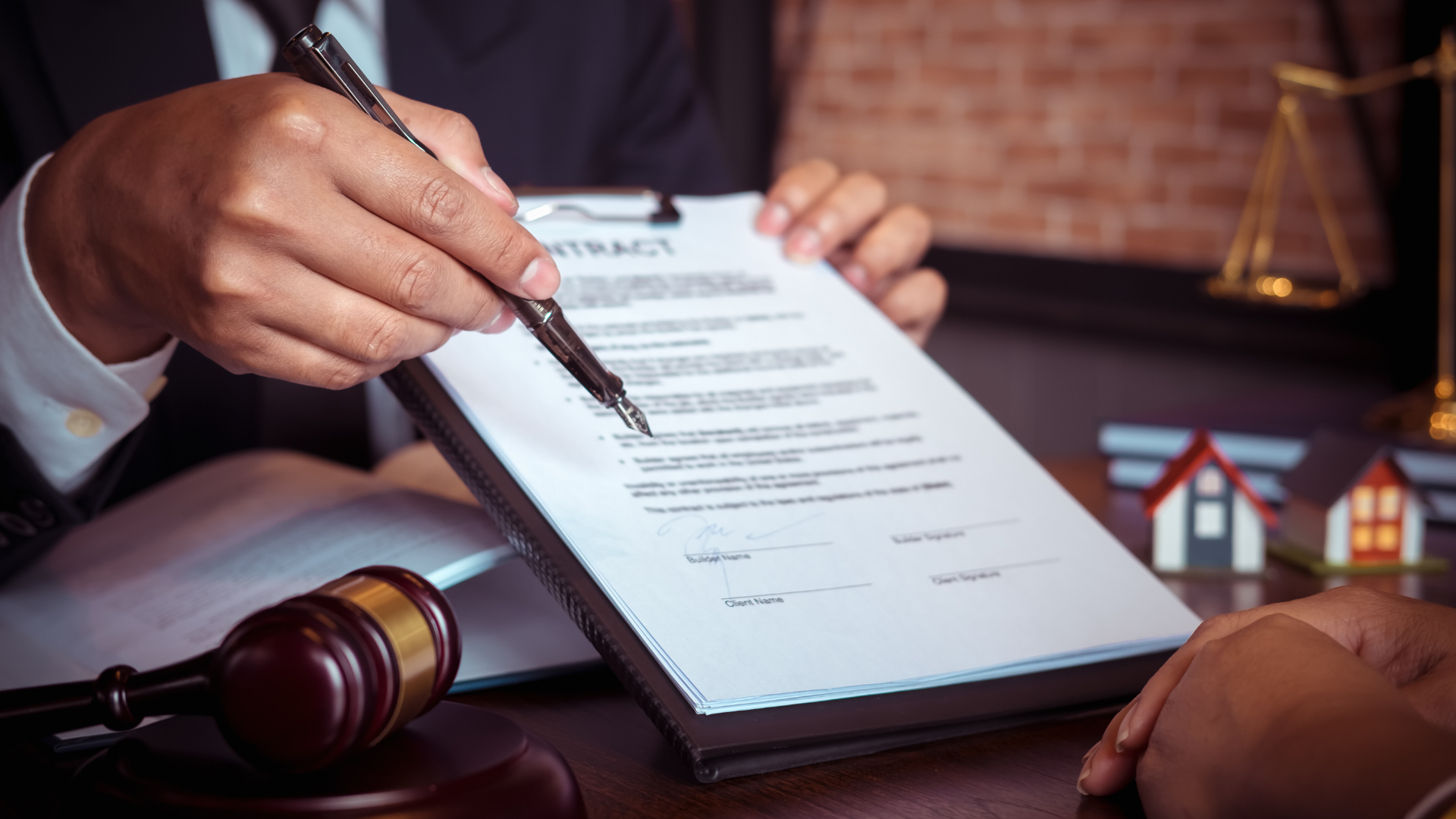 Person in suit points to a contract on a clipboard; gavel, scales, and miniature houses sit nearby.