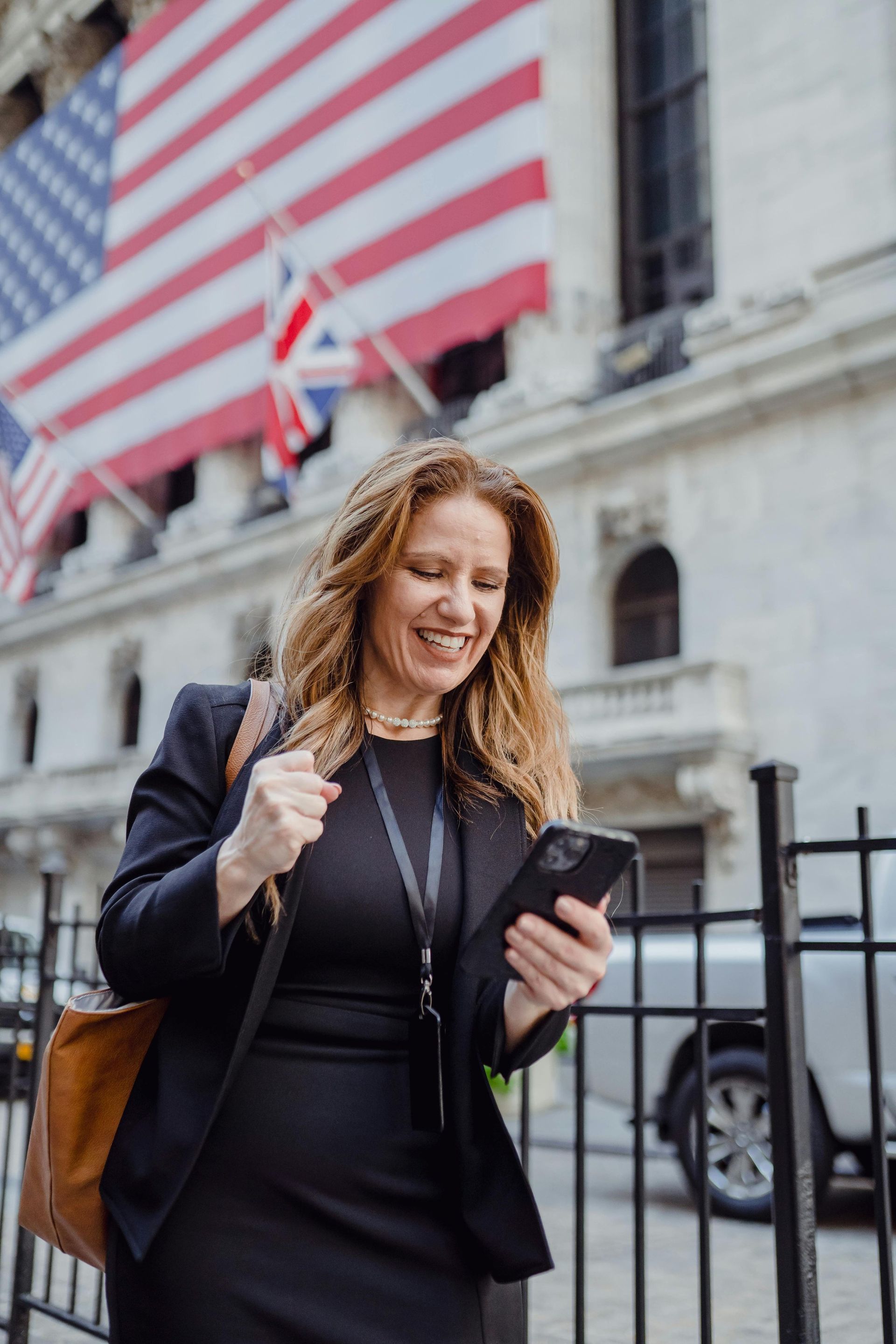 Woman in black suit smiles at phone, outside a building with American and British flags.