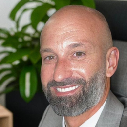 Bald man with a gray beard smiling, wearing a gray suit. Green plant in background.