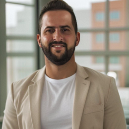 Man in tan blazer and white shirt smiles in front of a window.