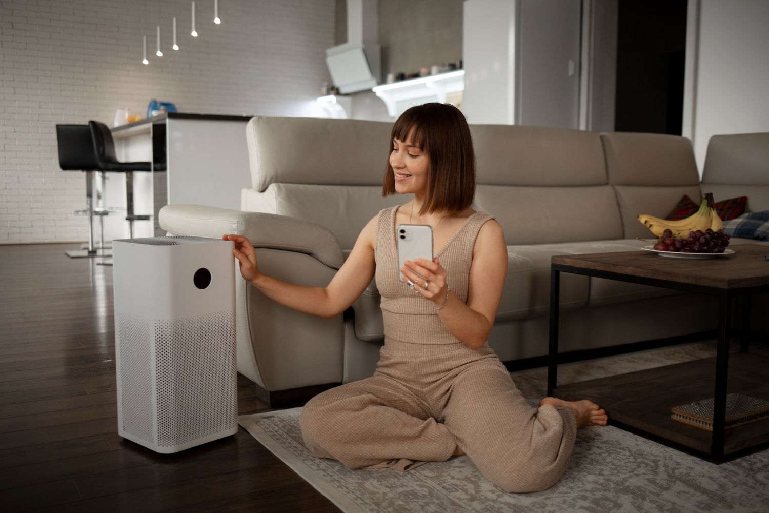 Woman kneels in a living room, using a smartphone beside a white air purifier near a couch.