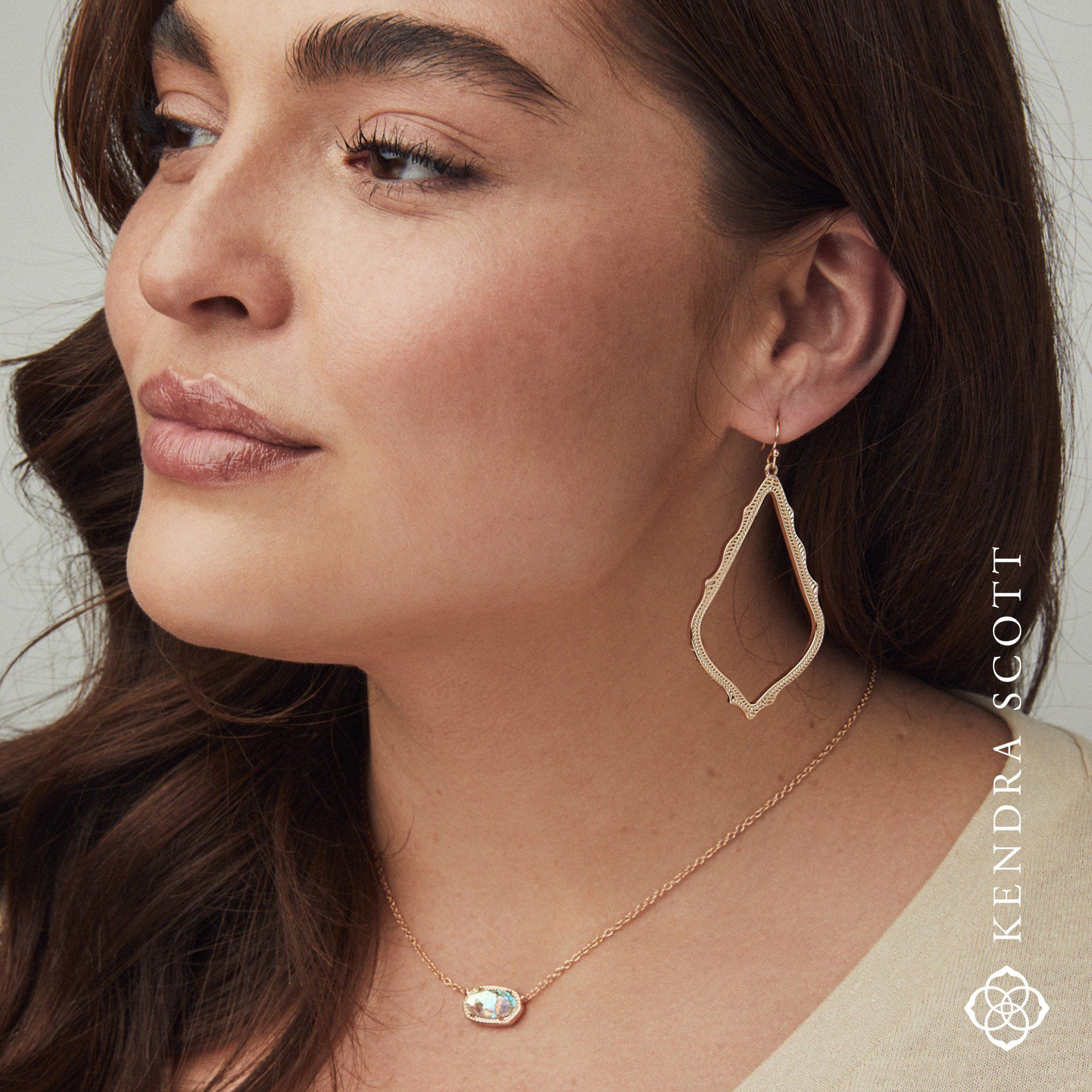 Woman wearing gold teardrop earrings and a necklace; neutral expression.