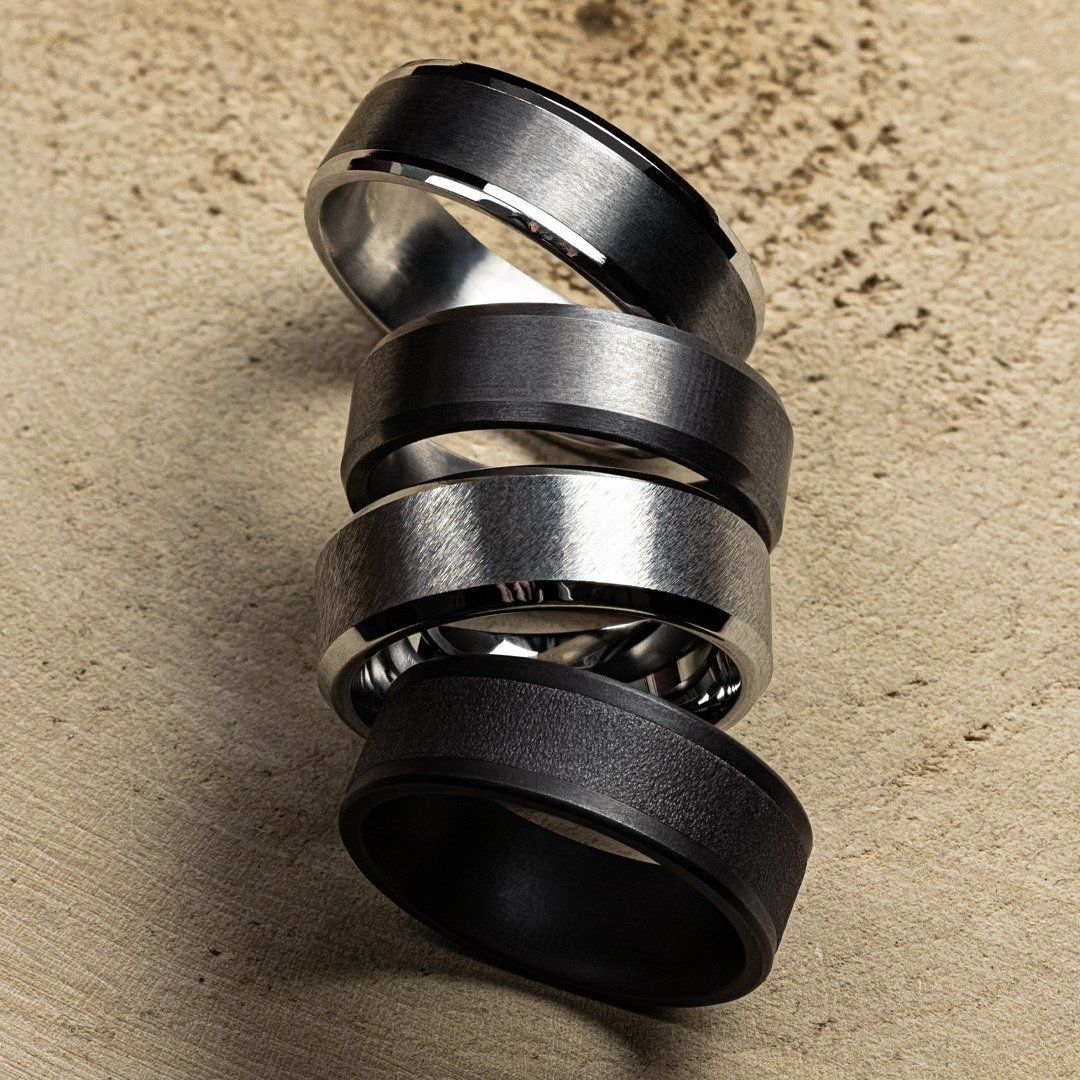 Stack of five men's rings: silver and black metal bands with various textures, on a wooden surface.