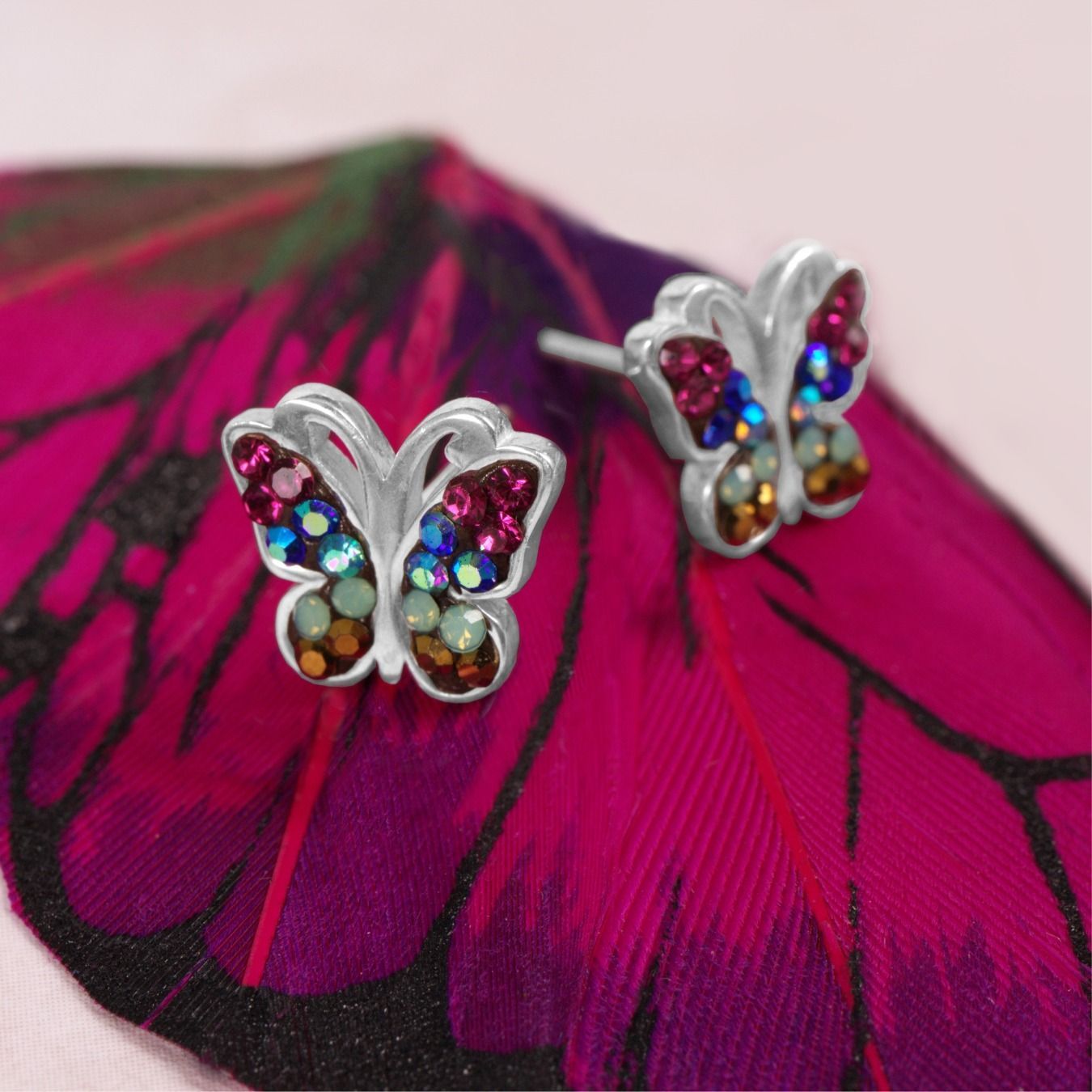 Butterfly stud earrings with rainbow-colored gems on a pink feather.