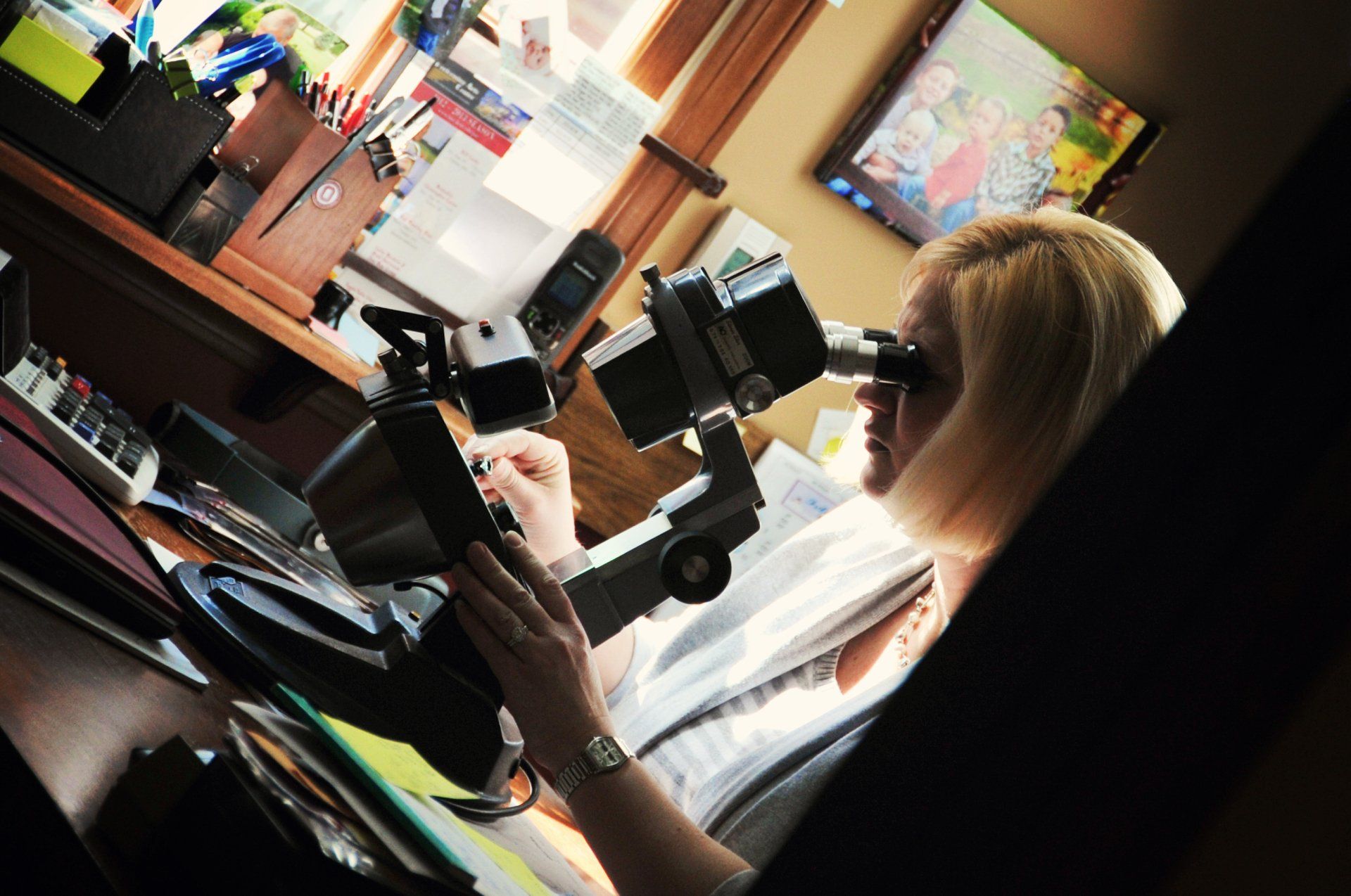 Woman looking through a microscope at a desk with papers. Blond hair, office setting, bright colors.