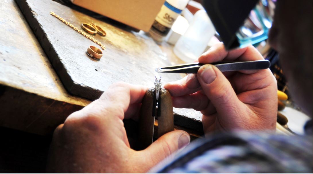 Jeweler using tweezers to work on a small piece of jewelry at a workbench.