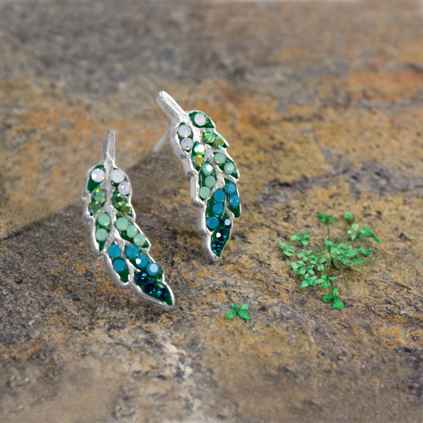Silver leaf-shaped earrings with green and blue gems on a textured stone surface.