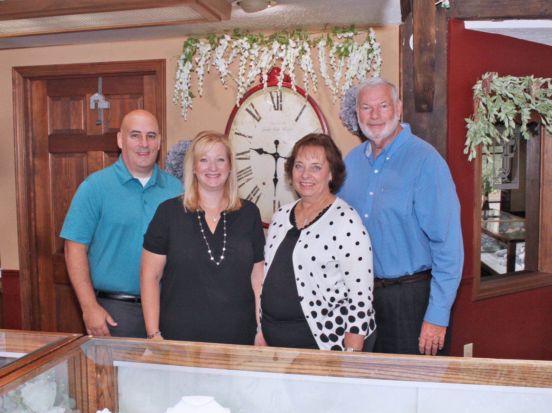 Four people standing behind a display case, possibly in a shop. Clock and flowers decorate the wall.