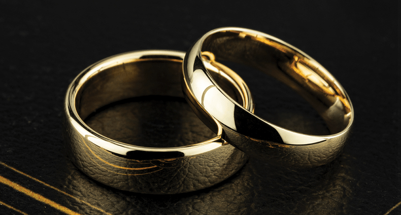 Two gold wedding rings, one with a subtle etched design, on a dark textured surface.