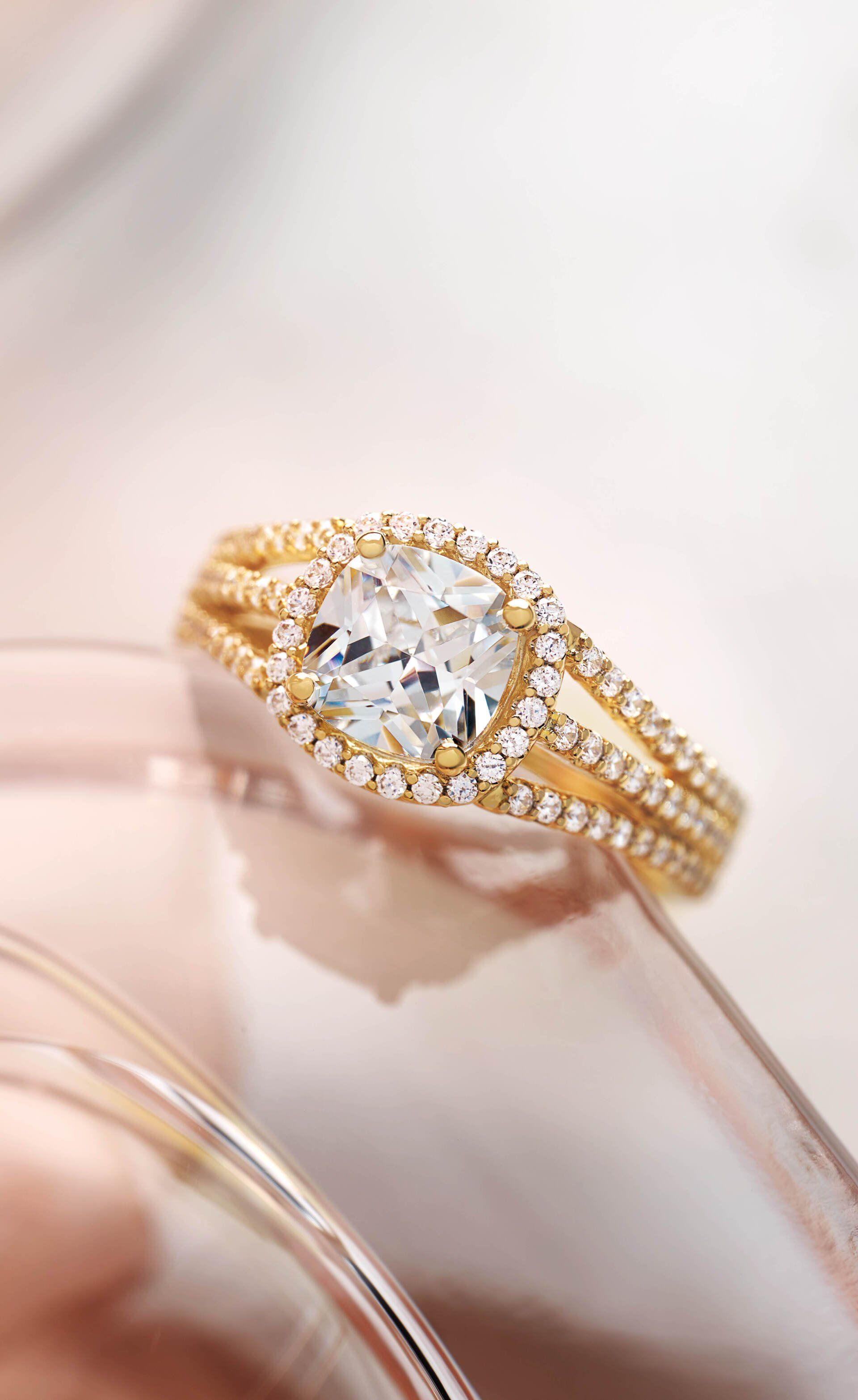 Gold ring with a large, square, clear gemstone surrounded by a halo of smaller stones; set on a reflective surface.
