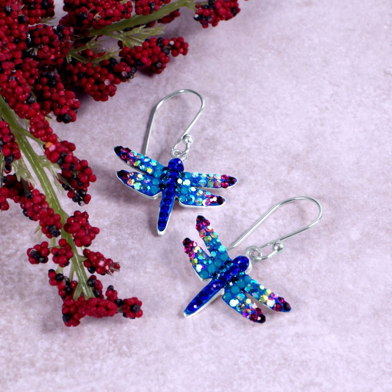 Dragonfly earrings with blue and purple crystal wings, on a pink surface next to red berries.
