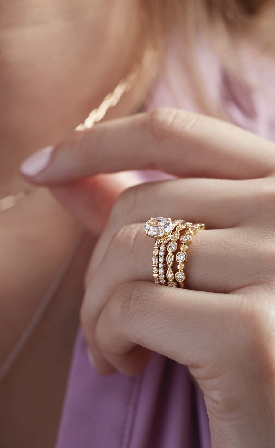 Woman's hand with gold rings, including a diamond engagement ring and stacked bands, touches a necklace.