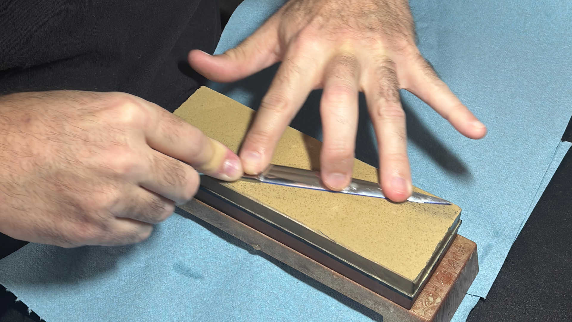 Hands sharpening a blade on a whetstone. The blade moves across the stone with pressure from the fingers.