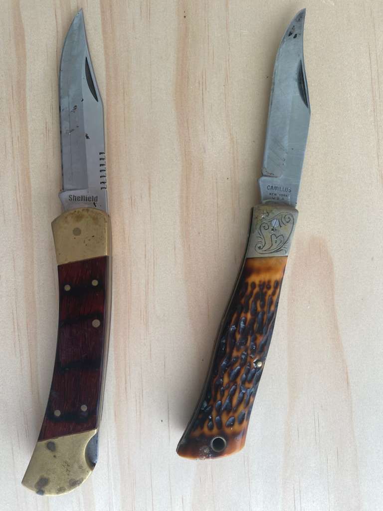Two folding knives on wooden surface; one with red handle, one with brown.