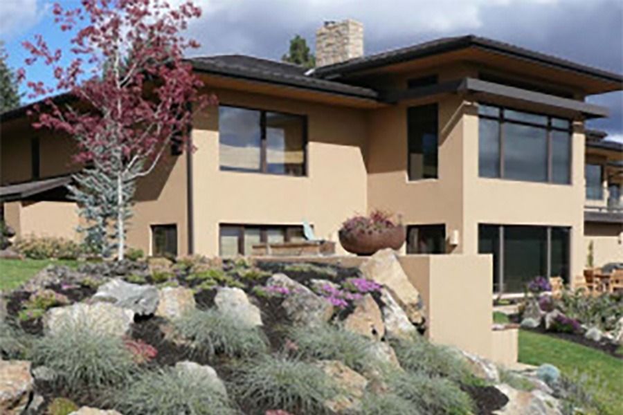 Modern tan house with landscaped garden, flowering tree, and rocky slope under a clear sky
