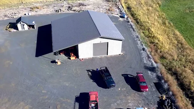 Aerial view of a gray metal building with parked vehicles on a gravel lot beside fields