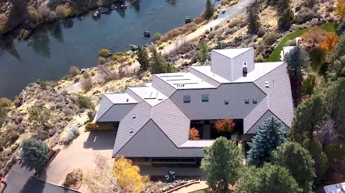 Aerial view of a tan house with a gray roof beside a river and wooded hillside.