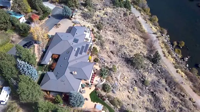Aerial view of a gray-roofed house beside a rocky hillside and riverbank.