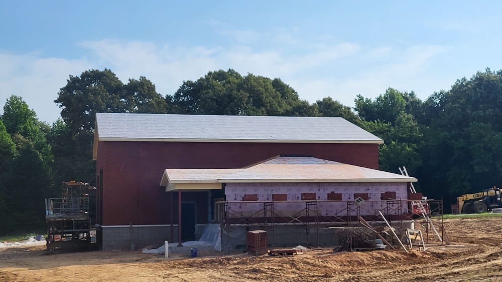 Red building under construction with a smaller, covered entrance. Trees and blue sky in the background.