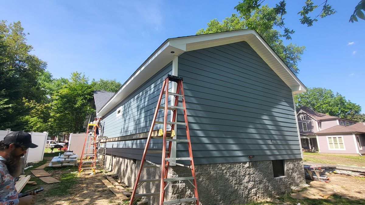Construction workers install blue siding on a house with a stone foundation under a blue sky.