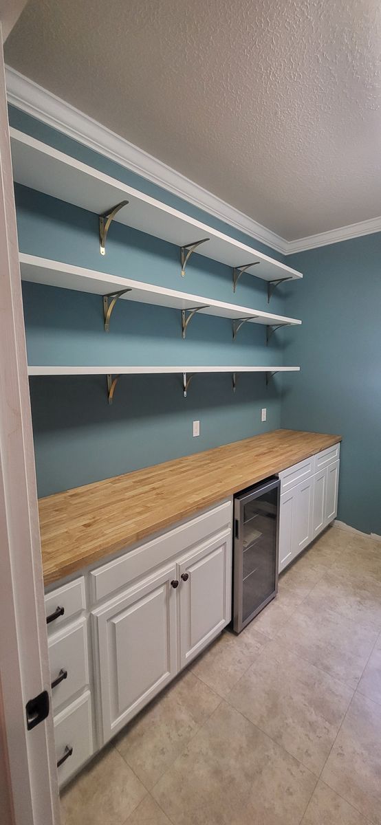 A pantry with white cabinets, wooden countertop, shelves, and a built-in wine fridge. The wall is blue.