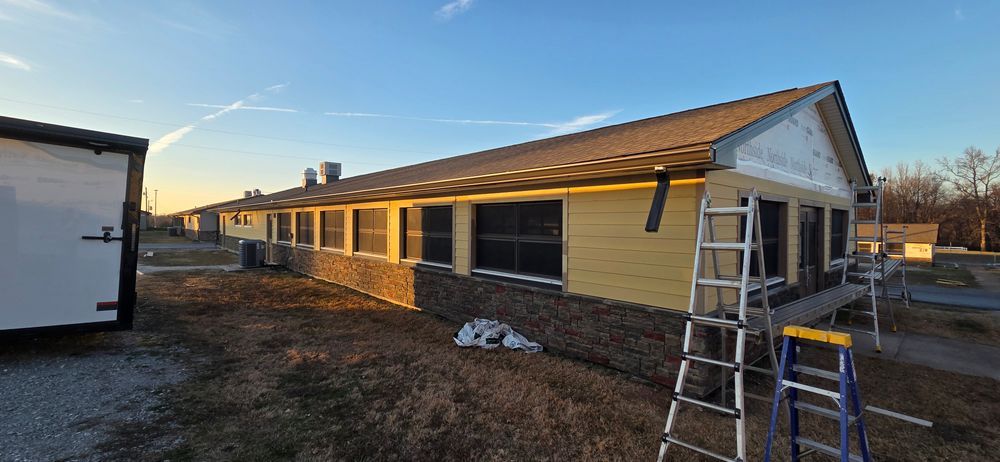 Exterior of a building under renovation with siding, ladders, and a trailer. Brown and yellow tones.