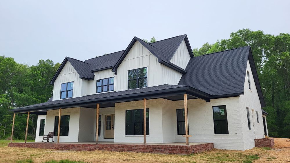 White farmhouse with a black metal roof and trim, brick foundation, and wraparound porch.