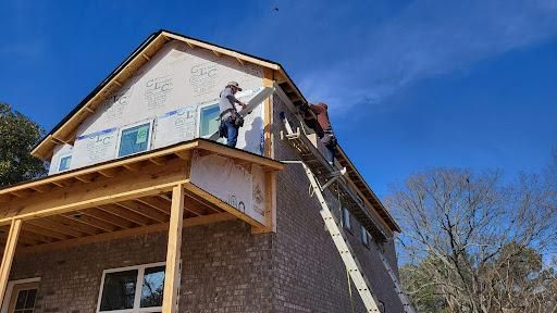 Construction workers installing siding on a two-story house, using a ladder on a sunny day.