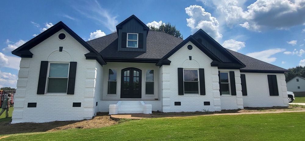 White brick house with black trim, shutters, and roof under a cloudy blue sky.