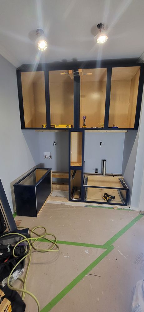 Kitchen cabinets during installation; black frames with light wood interiors. Construction tape on floor.