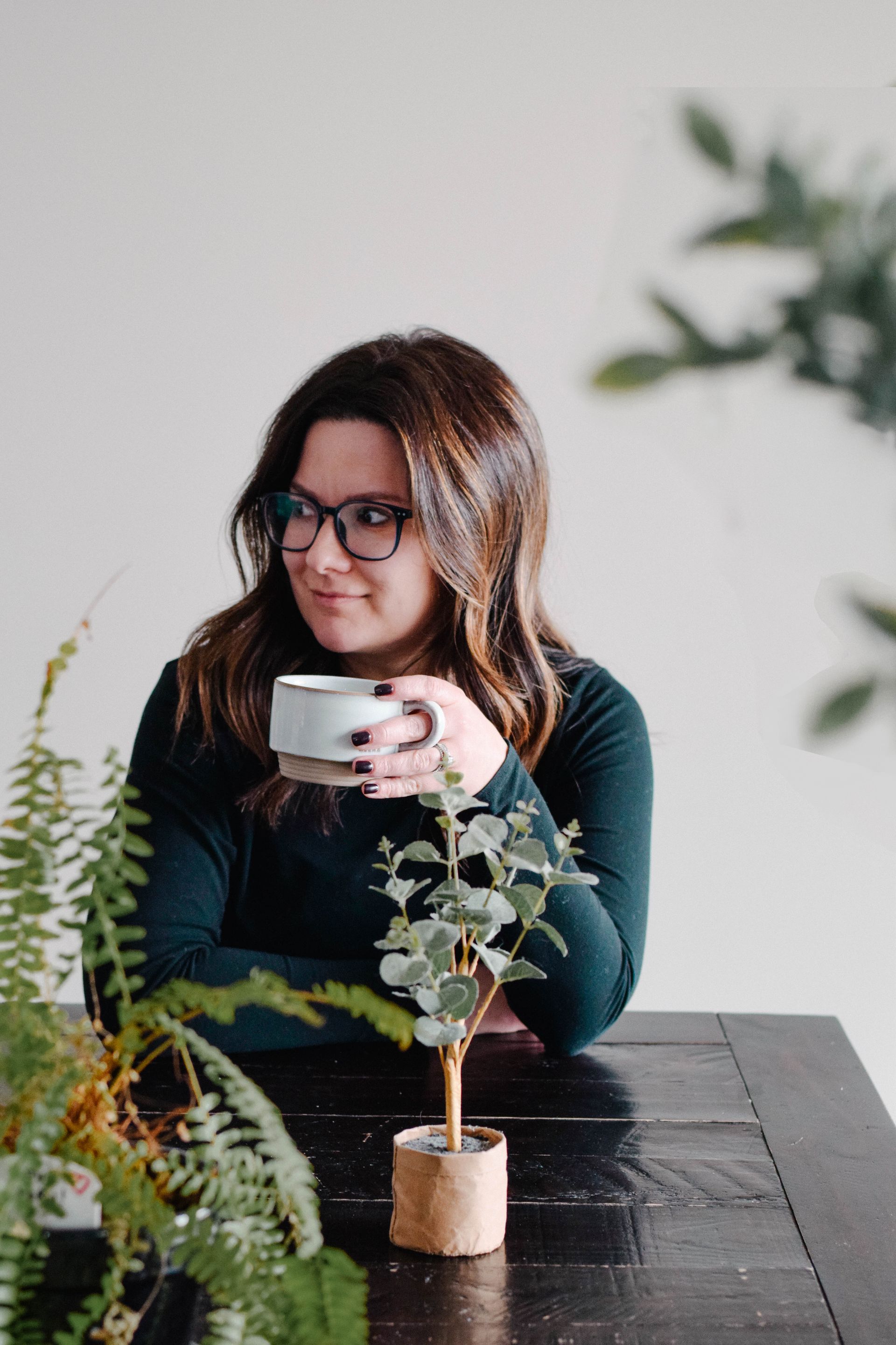 Woman with glasses, holding a mug, sitting at a dark table. Small potted plant and green foliage in the foreground.