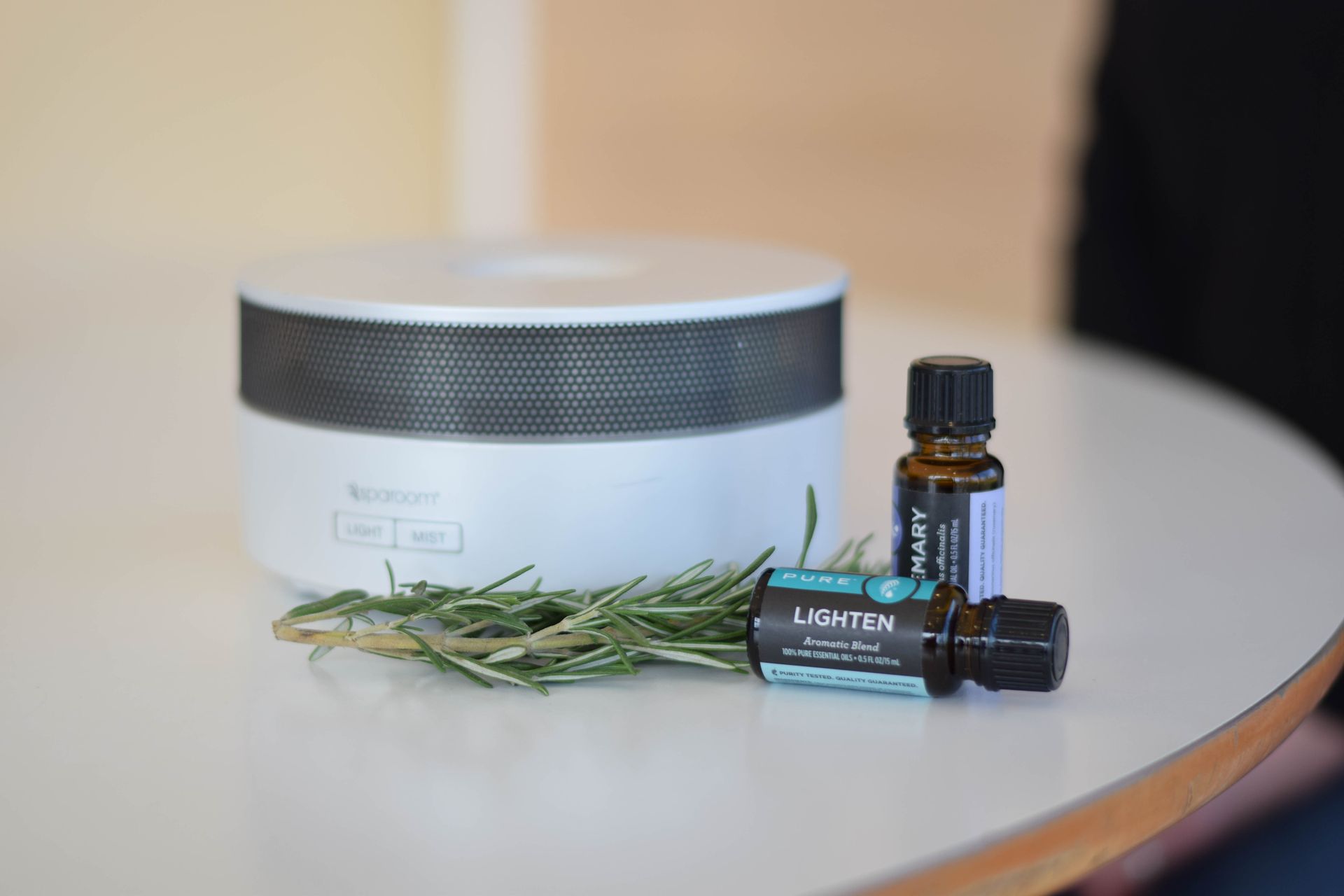 White essential oil diffuser, two oil bottles, and rosemary sprig on a white table.