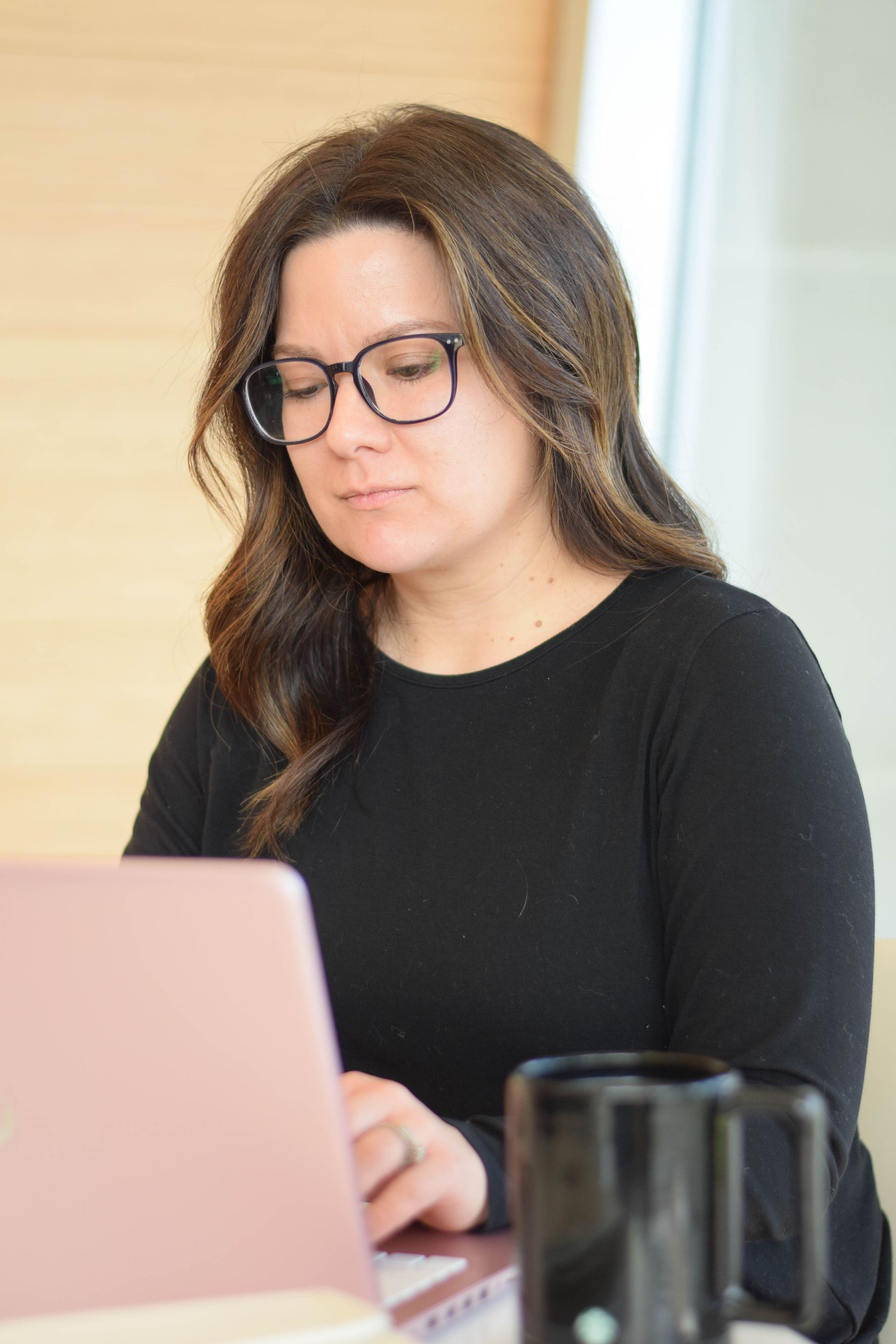 Woman with glasses working on a pink laptop at a table, black mug nearby.