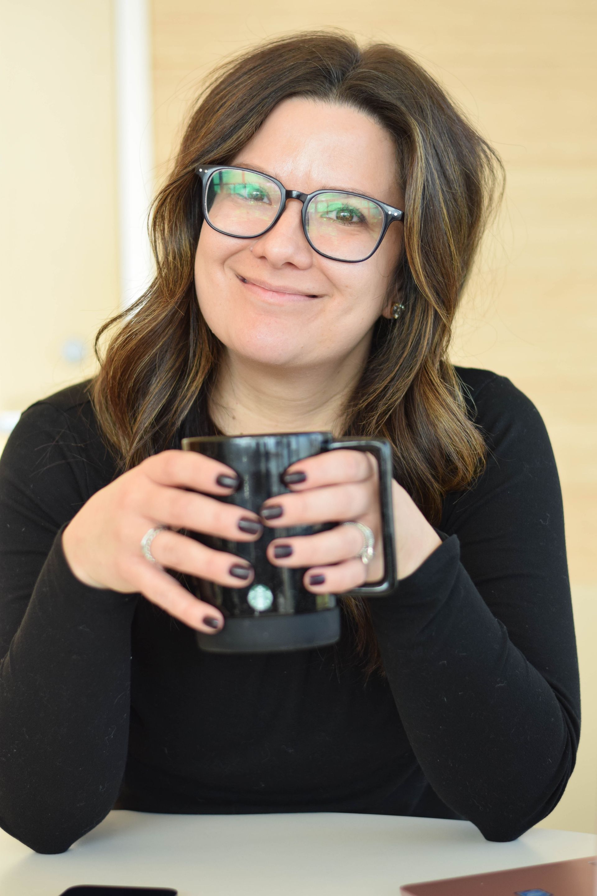 Woman with glasses smiles, holding a black mug. She wears a black top, sitting indoors.