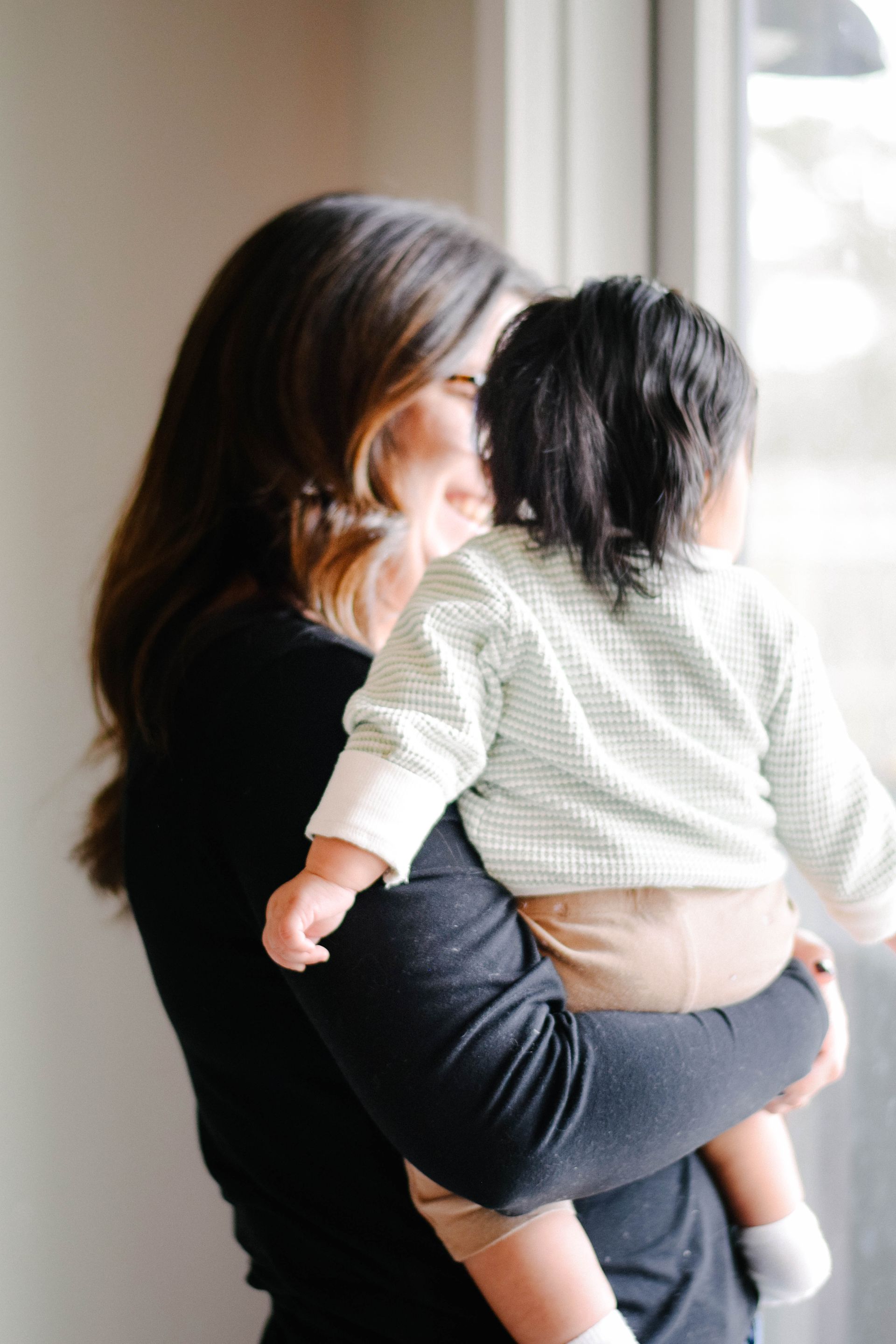 Woman holding a baby, looking out a window. Baby in light green striped shirt, facing window.