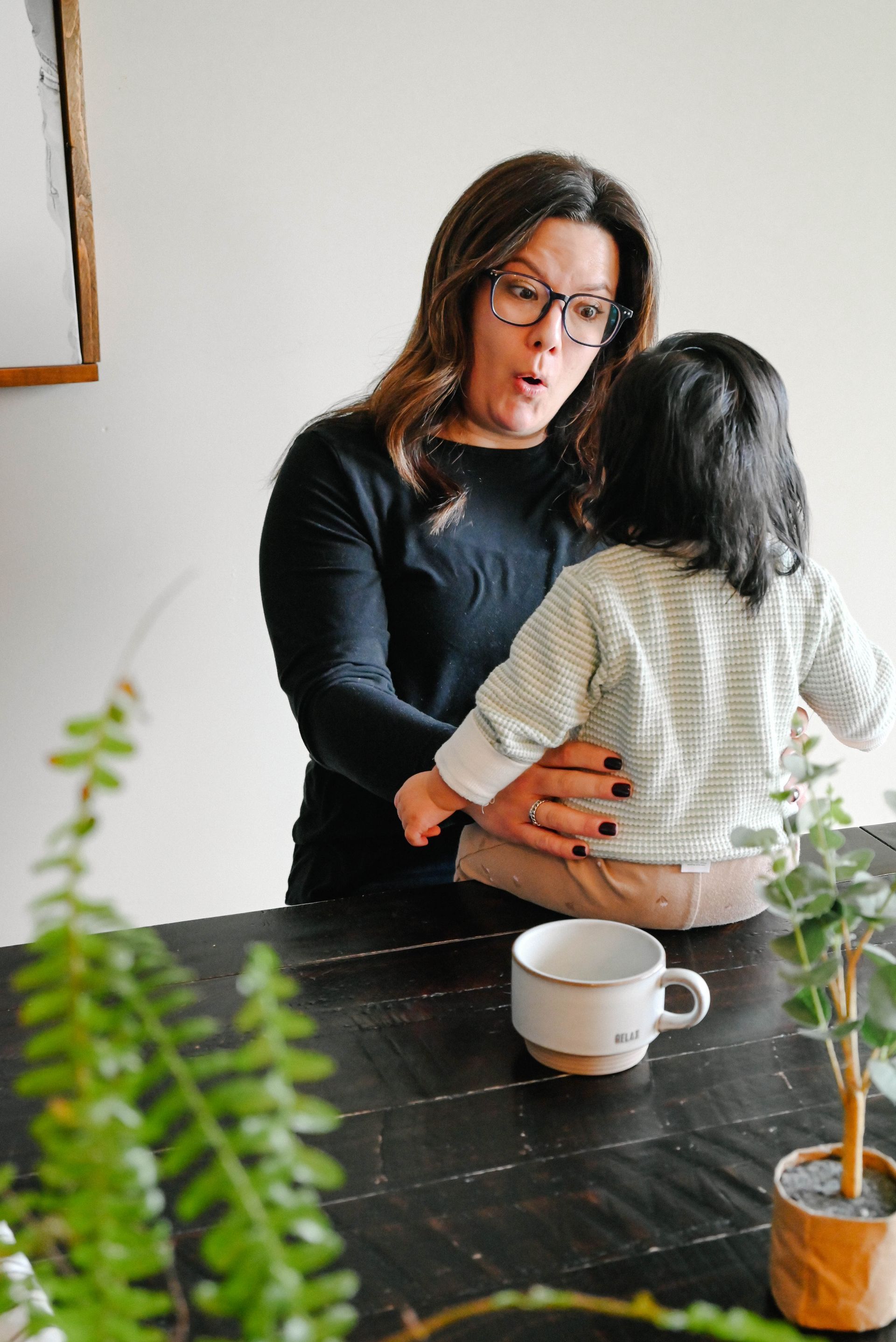 Woman with glasses talking to a child sitting on a table, coffee cup present, indoor setting.