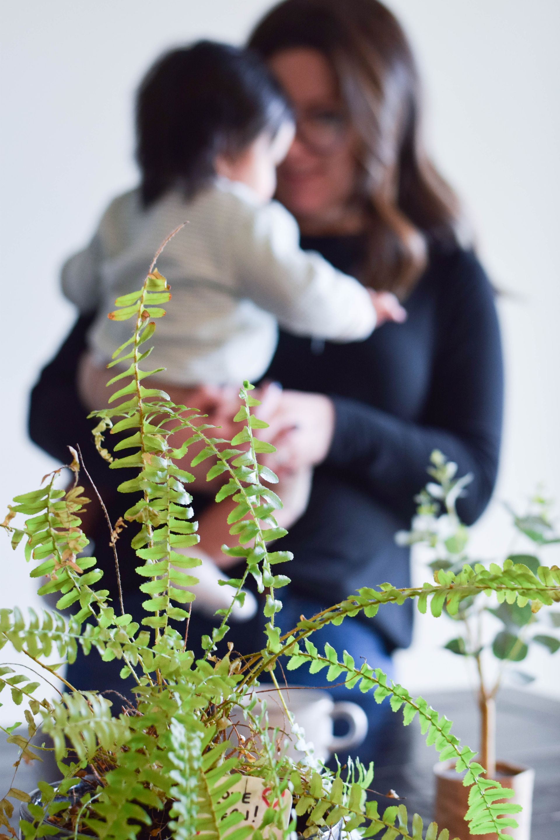 Woman holding a baby, in front of leafy plants.