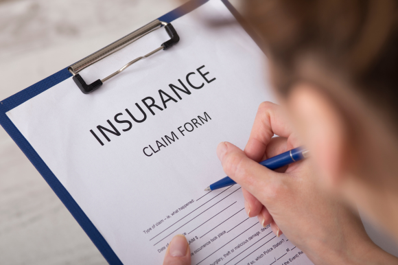 Person writing on an insurance claim form, blue pen in hand.