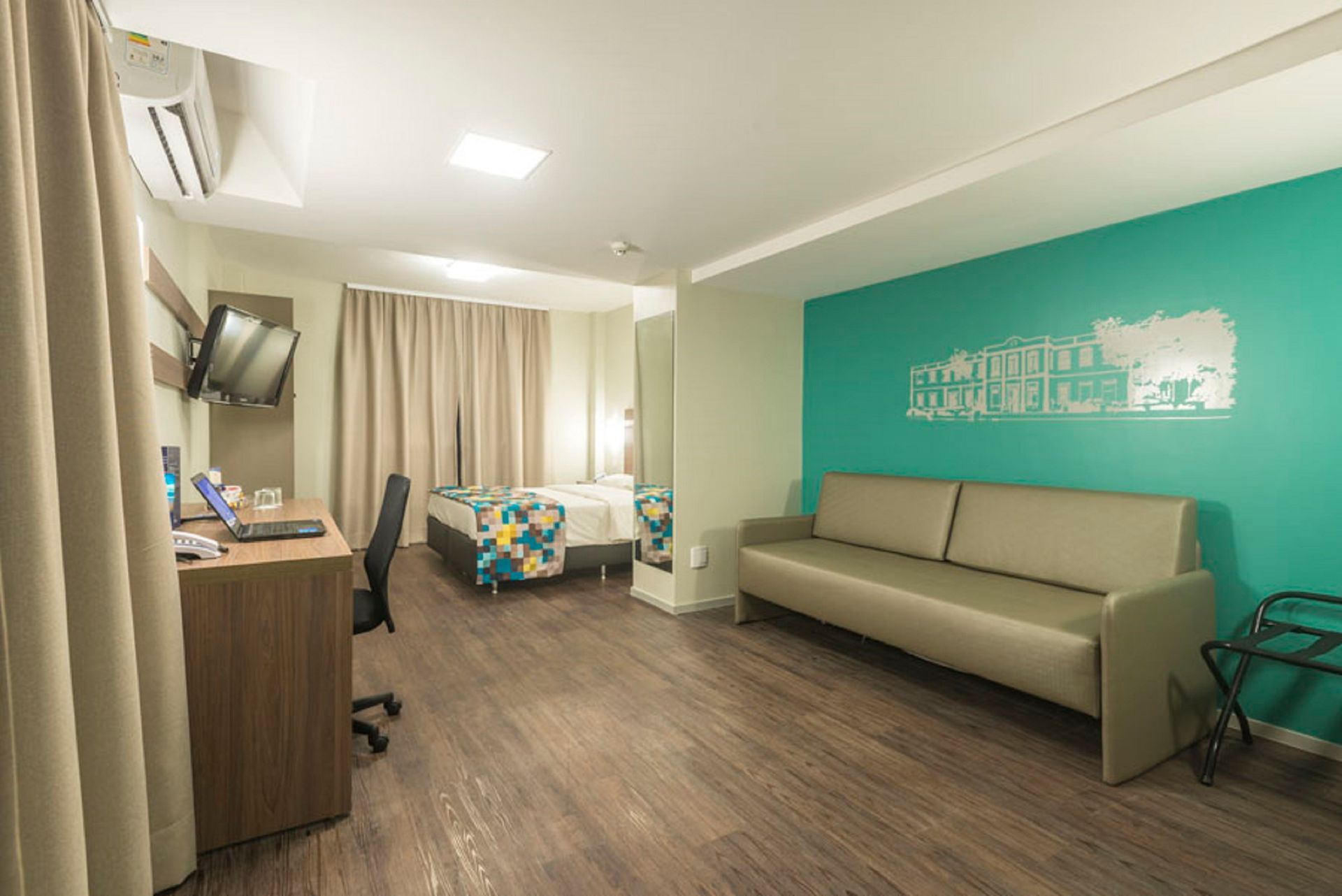 Quarto de hotel moderno com cama, escrivaninha, sofá e parede com detalhes em turquesa.