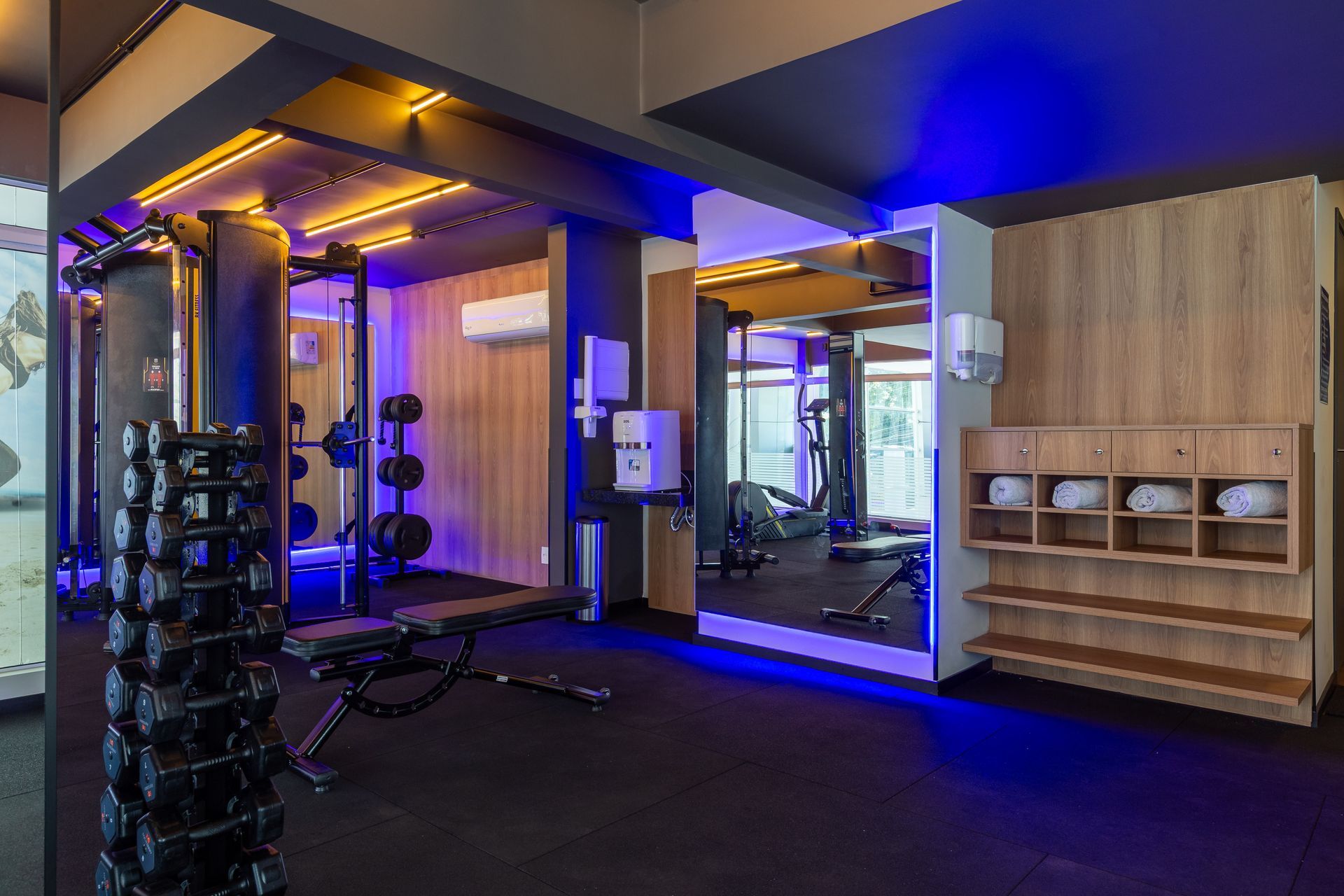 Interior de academia moderna com equipamentos de ginástica, espelhos, iluminação azul e prateleiras de armazenamento com painéis de madeira.