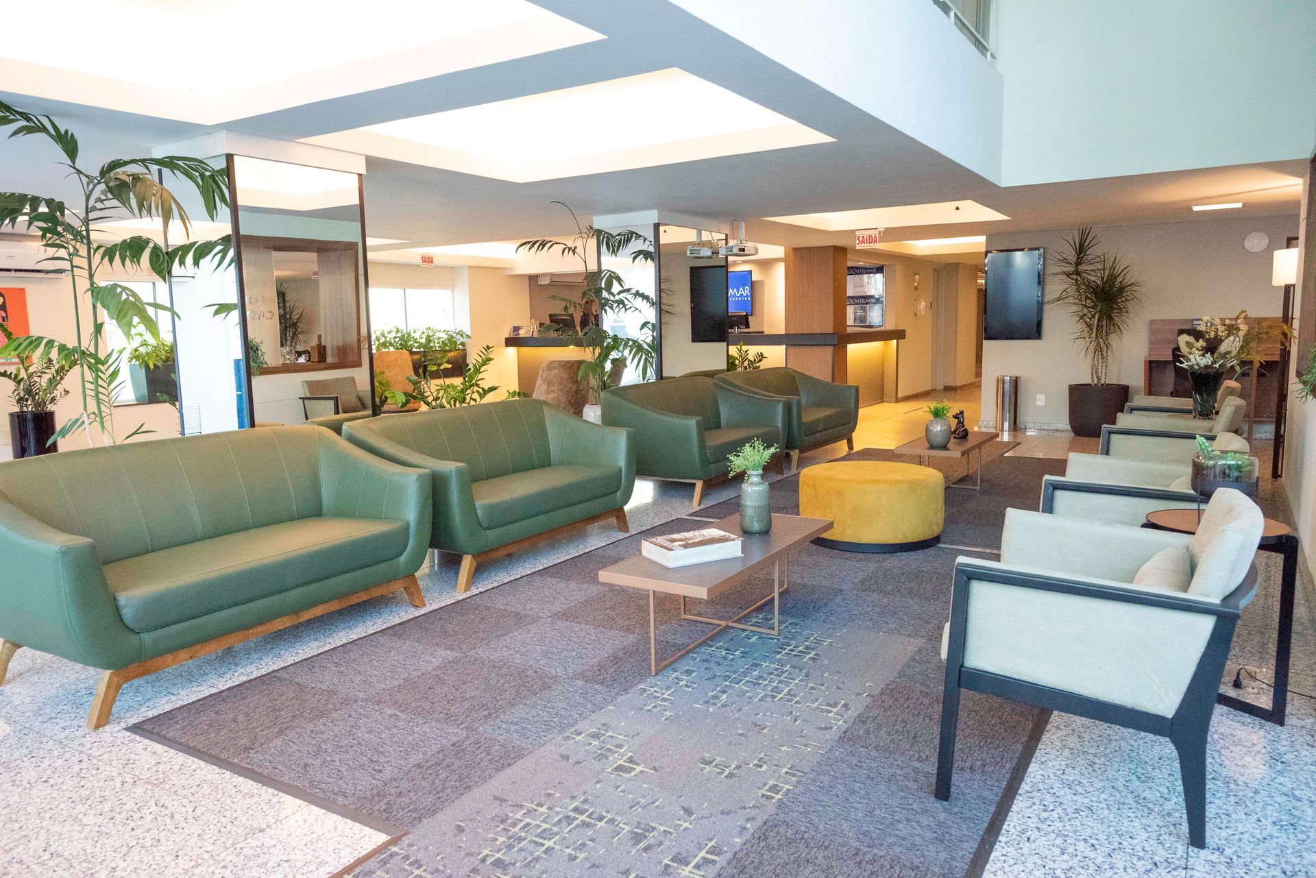 Sala de estar moderna com sofás verdes, poltronas brancas, pufe amarelo e plantas sob luzes brilhantes no teto.