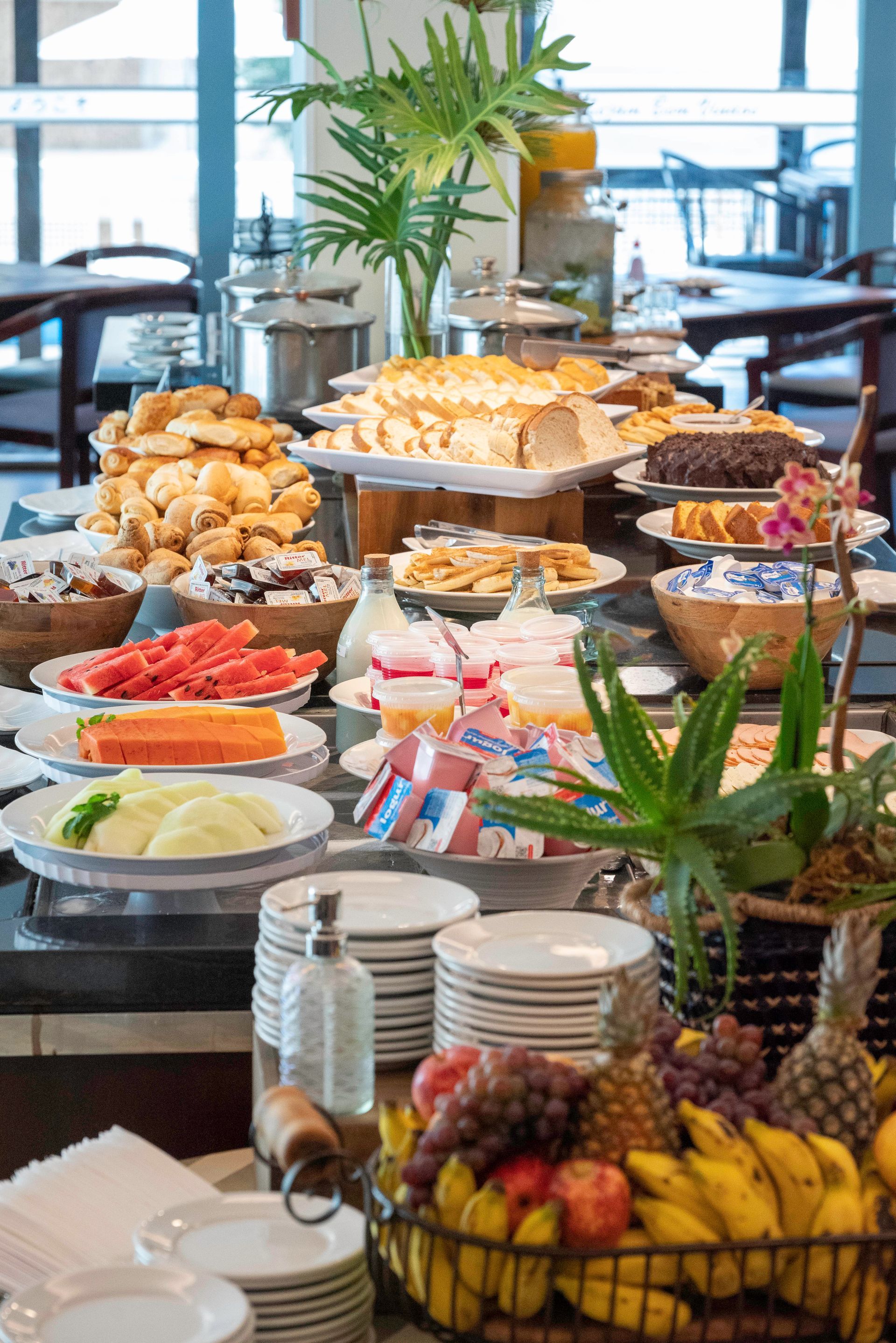 Mesa de buffet com uma variedade de doces, frutas e café em uma sala de jantar iluminada.