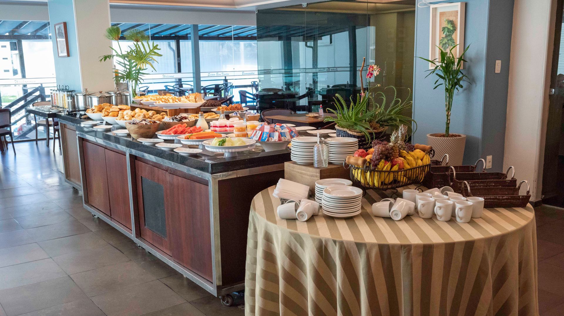 Mesa de buffet com doces, frutas e pratos empilhados em uma área de jantar iluminada.