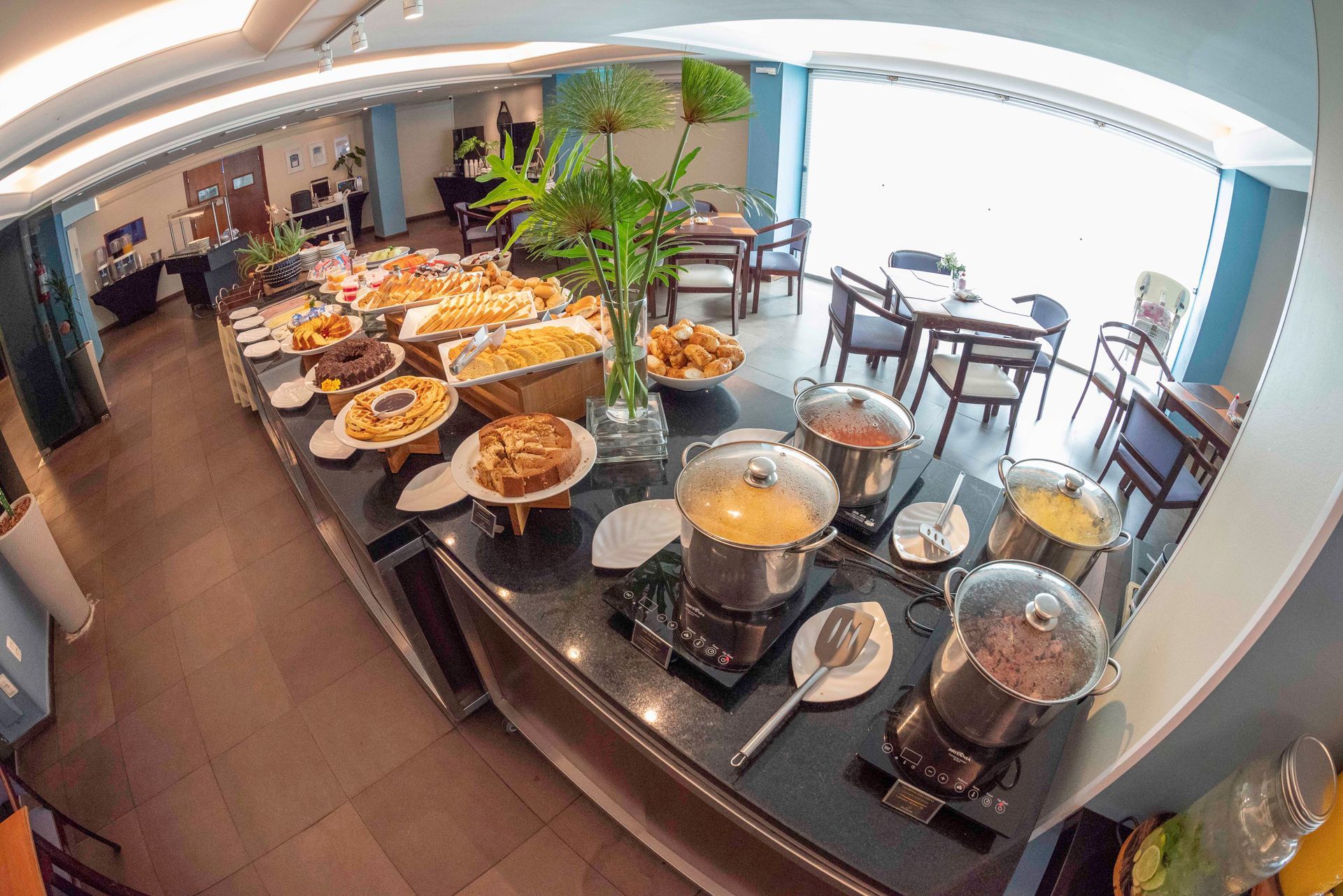 Buffet de café da manhã com doces, frutas e pratos cobertos em um balcão em uma área de refeições iluminada.
