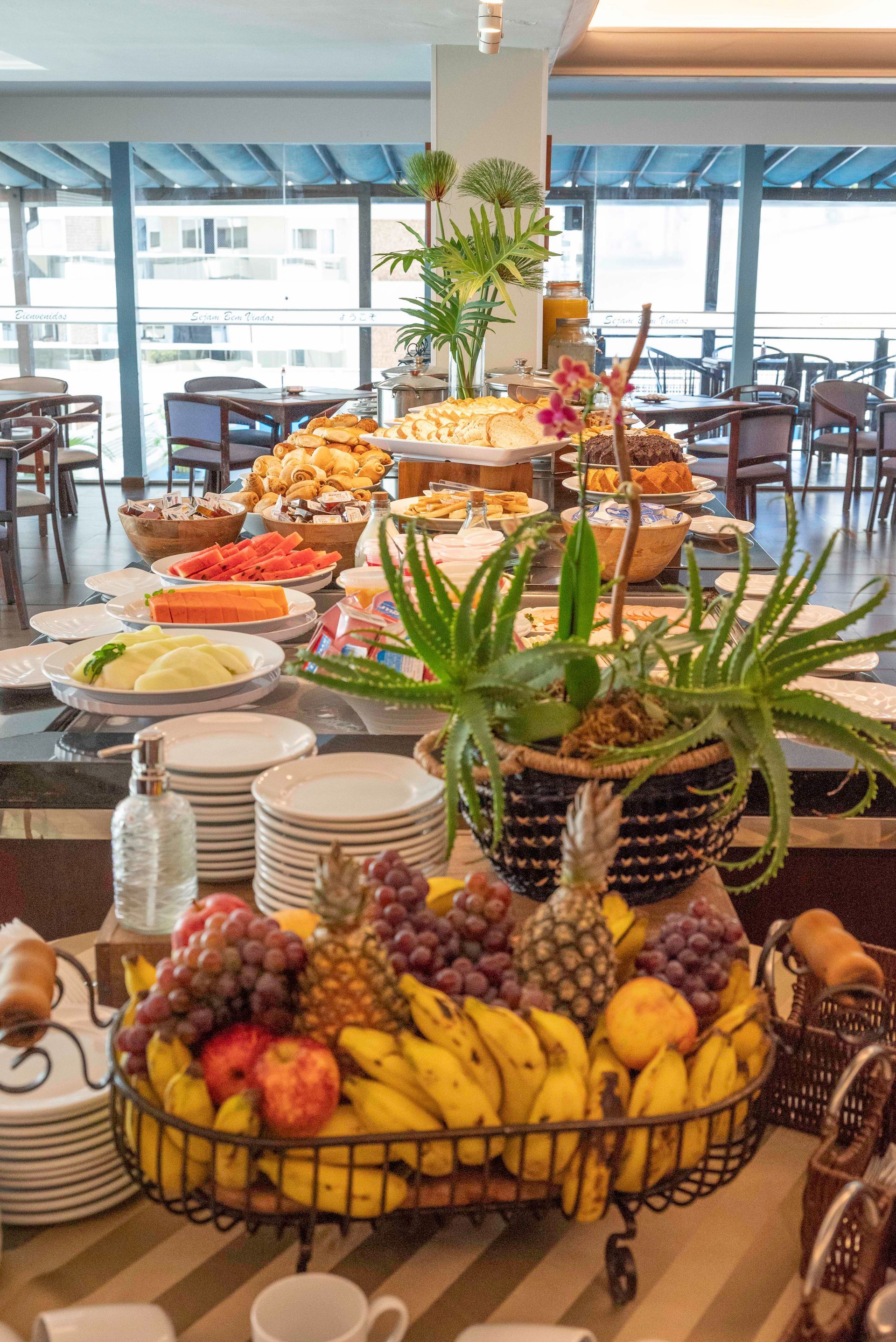 Mesa de buffet com travessas de frutas variadas em uma sala de jantar iluminada com mesas e cadeiras.
